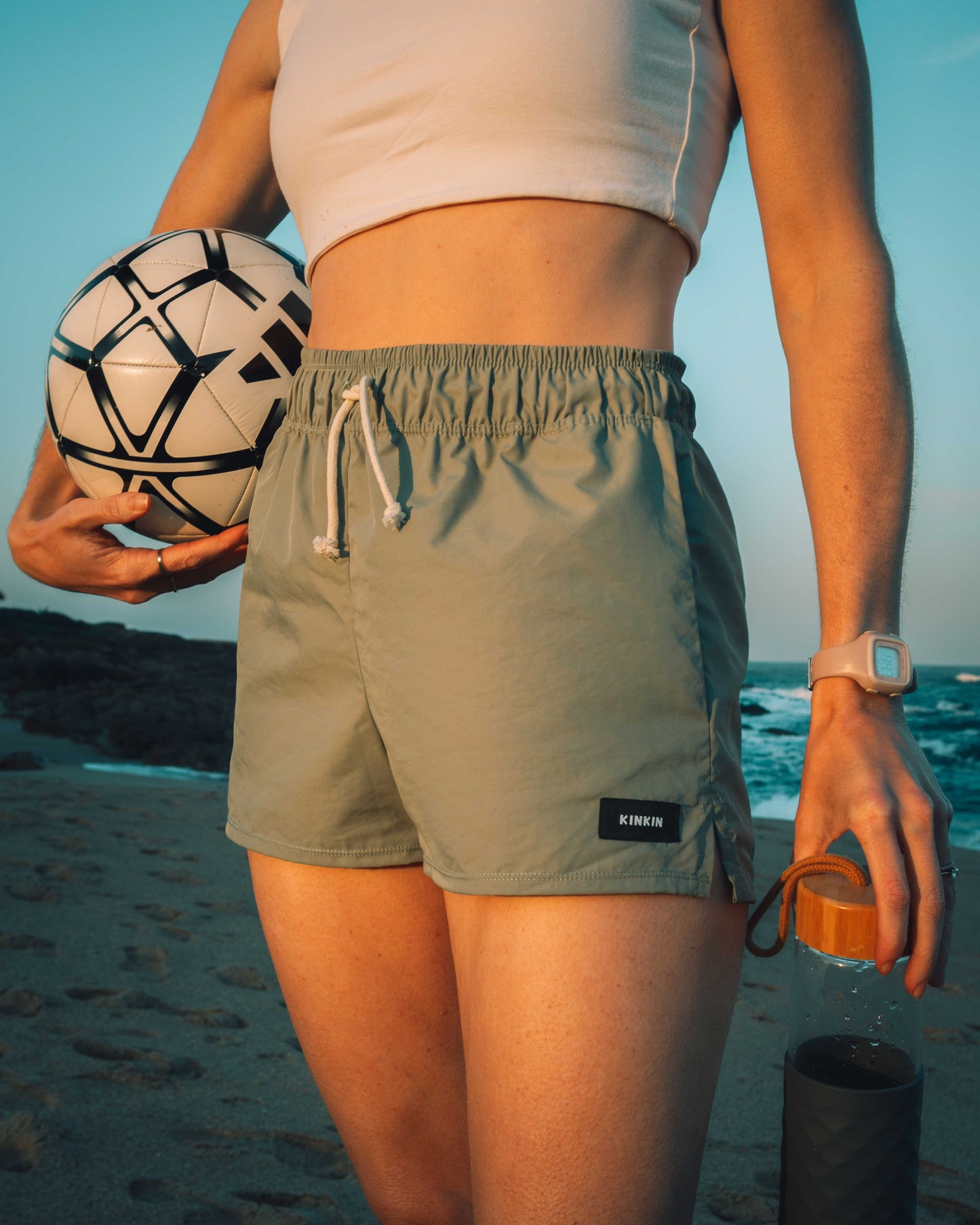 Person on a beach holding a volleyball and a drink, wearing green shorts with a brand logo.