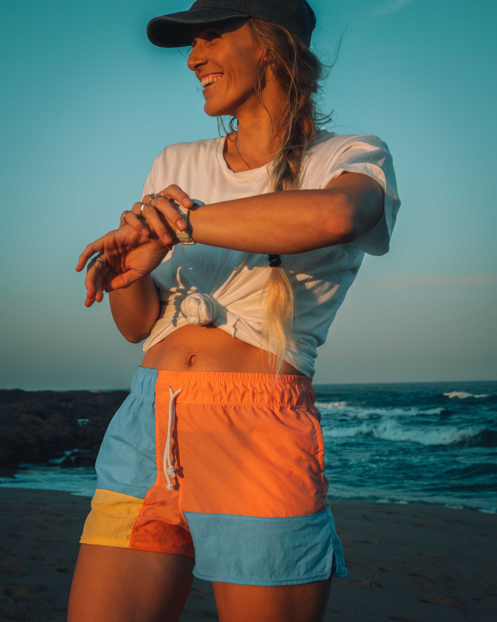Woman on a beach at sunset, wearing a white shirt and colorful shorts.