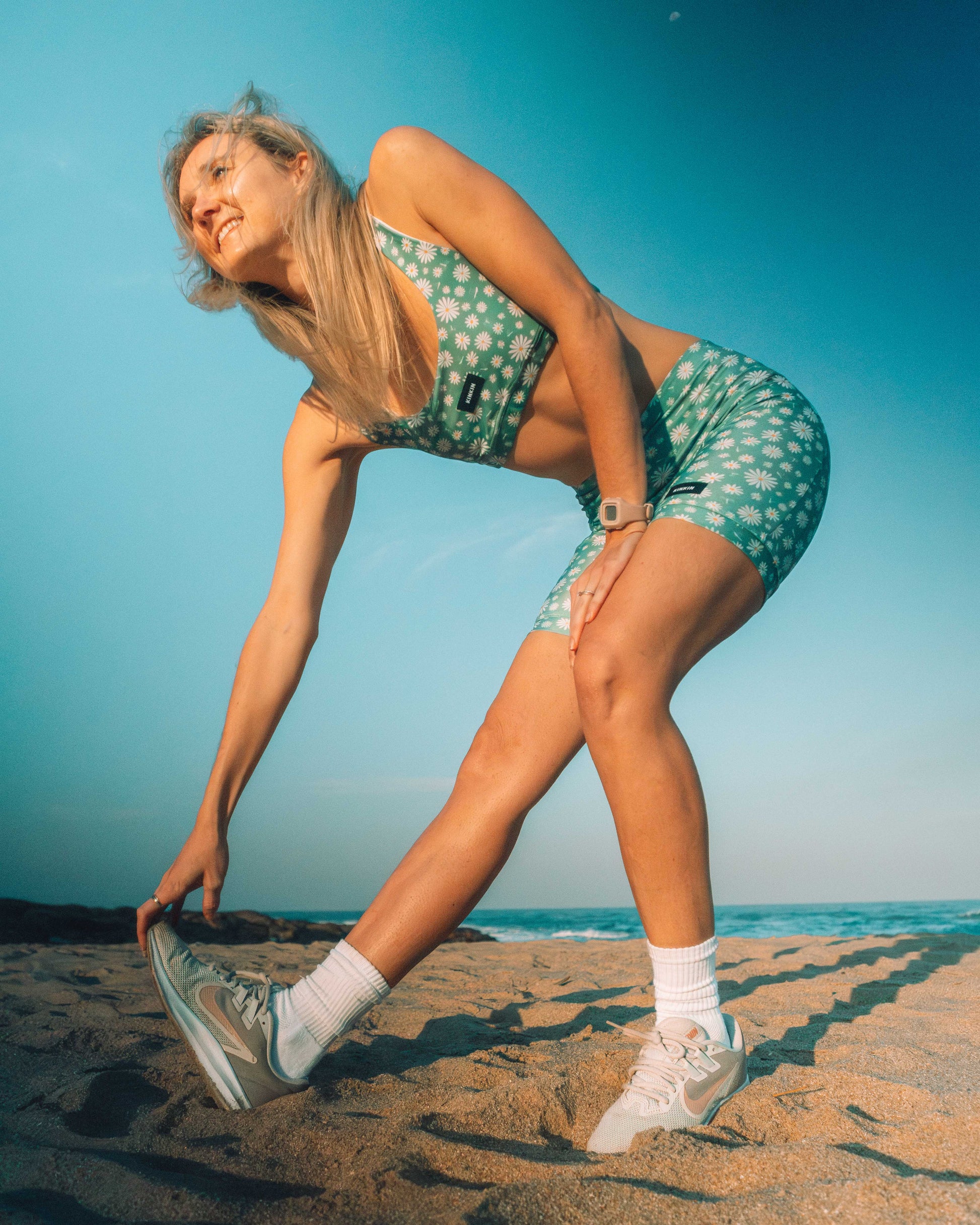 Woman in a green swimsuit stretching on a beach with a clear blue sky.